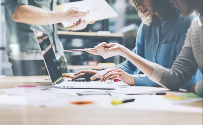 Zwei Personen arbeiten gemeinsam an einem Laptop, eine dritte Person reicht ein Dokument