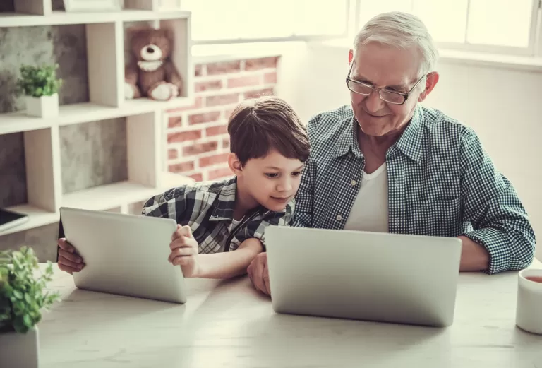 Ein Mann und ein Junge sitzen an einem Tisch mit vor ihnen geöffneten Laptops, drinnen. Die Umgebung umfasst Möbel und eine Wand im Hintergrund.