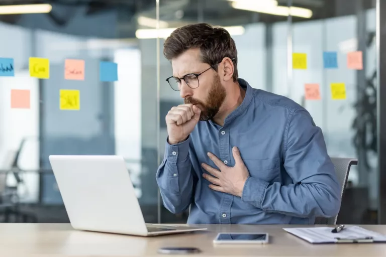 Ein junger männlicher Büroangestellter sitzt an einem Schreibtisch bei der Arbeit und hustet, bedeckt seinen Mund mit der Hand und hält seine Brust, atmet hart und fühlt sich unwohl.