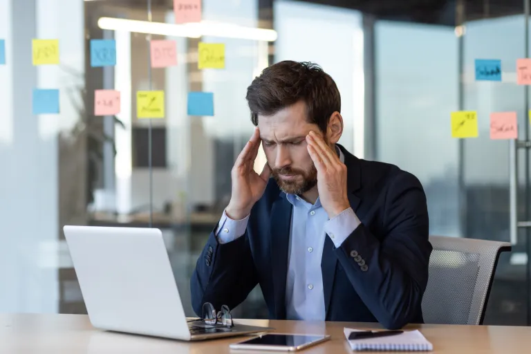 Ein gestresster Geschäftsmann, der am Schreibtisch sitzt und am Laptop arbeitet, den Kopf hält und Kopfschmerzen oder Frustration erfährt.
